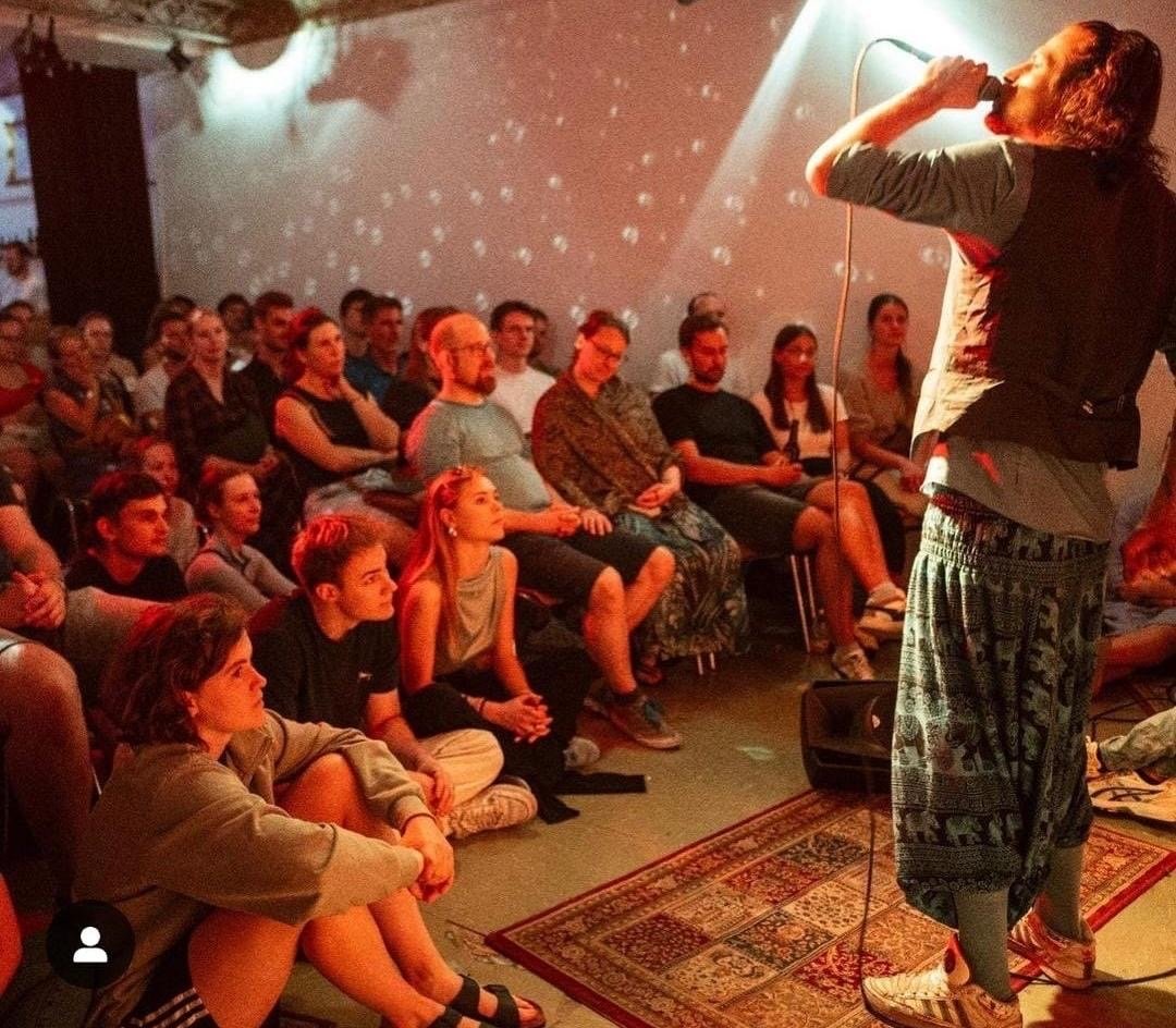 Ein Musiker mit langen Haaren, Weste und gemusterter Pluderhose rappt ins Mikrofon, vor ihm sitzt dicht gedrängt das Publikum auf Stühlen und auf dem Boden eines rot beleuchteten Raums mit Teppich und Lichtpunkten an der Wand.